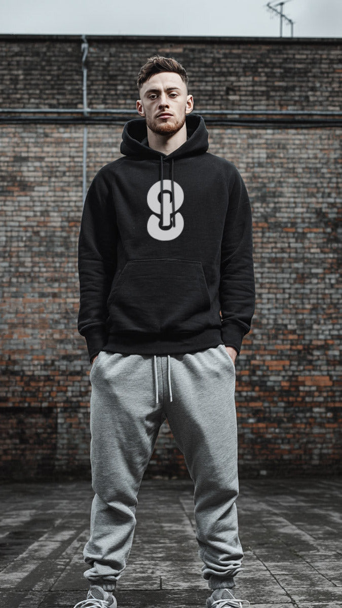 Man wearing a black hoodie with a logo and gray pants standing in front of a brick wall.