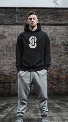 Man wearing a black hoodie with a logo and gray pants standing in front of a brick wall.