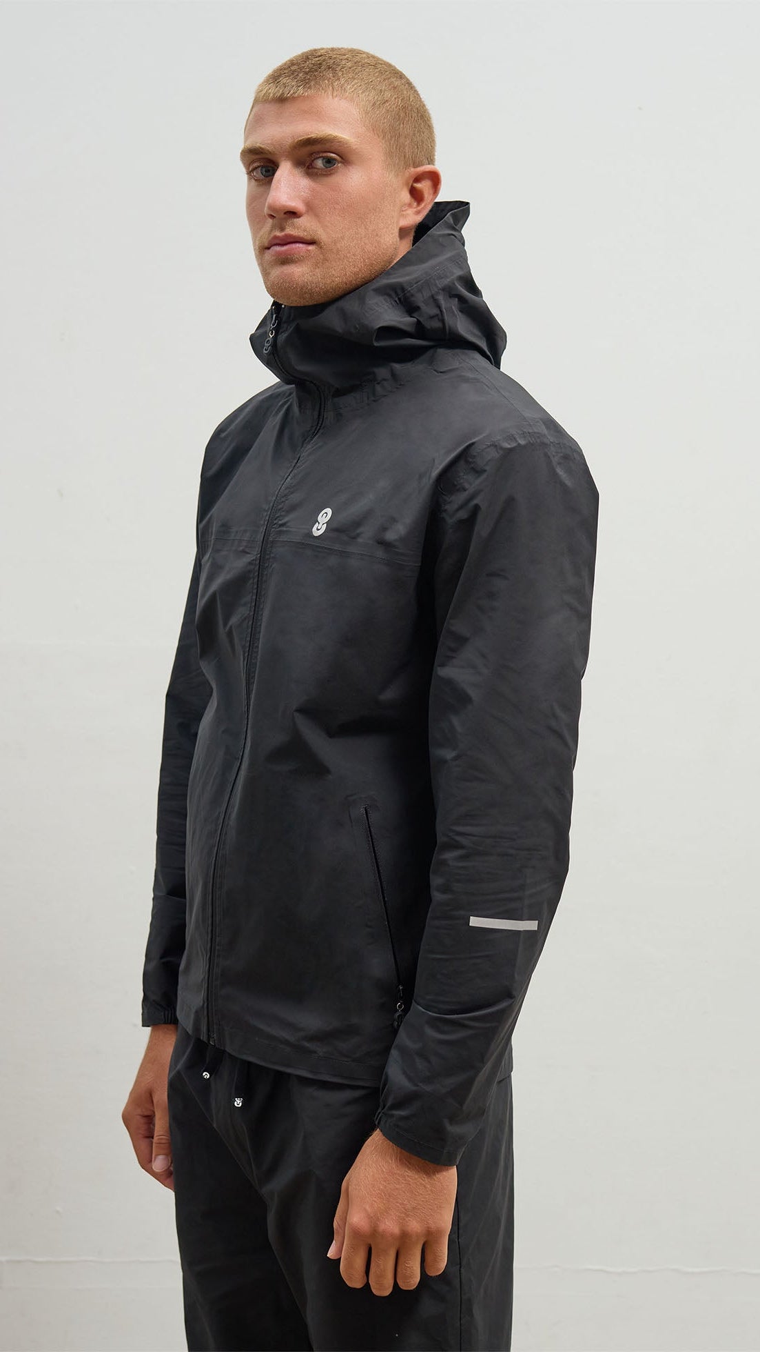 A person wearing a black OCL windbreaker jacket with a hood, featuring reflective branding and a zipper.