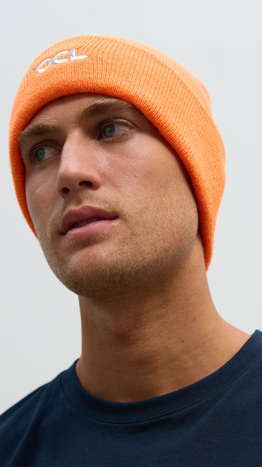 A person wearing an orange knit beanie with the OCL logo embroidered on the front.