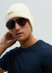 A person wearing a white knitted beanie and aviator sunglasses with the OCL logo embroidered in gold.