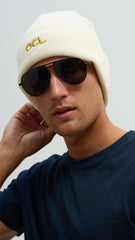 A person wearing a white knitted beanie and aviator sunglasses with the OCL logo embroidered in gold.