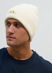A person wearing an off-white knitted beanie hat with the OCL logo embroidered in gold.