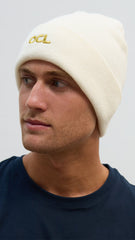 A person wearing an off-white knitted beanie hat with the OCL logo embroidered in gold.