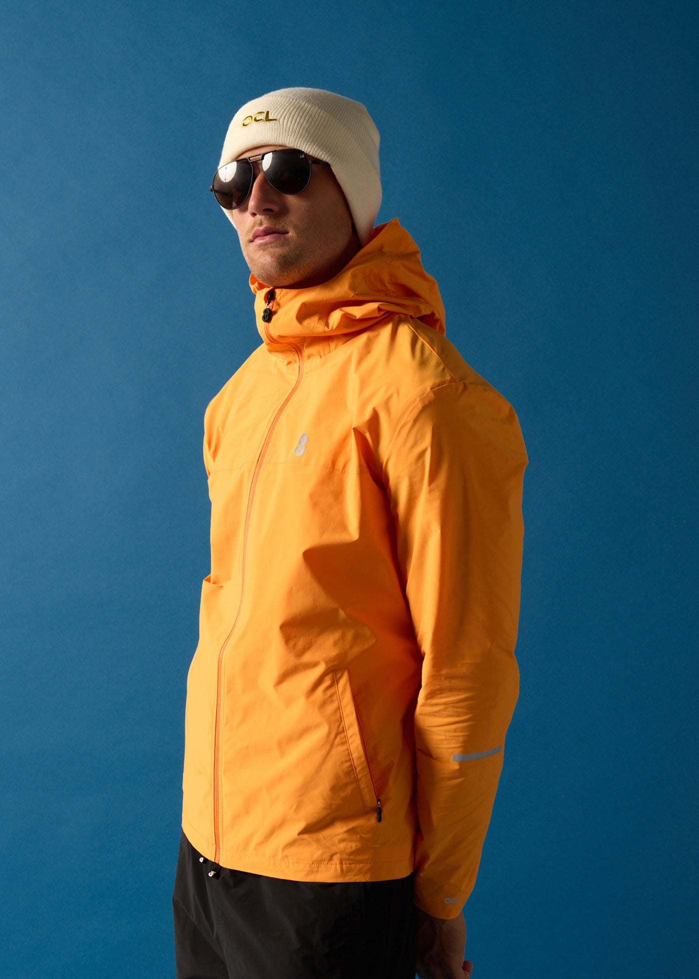 Person wearing an orange jacket, off-white OCL beanie with gold embroidery, and sunglasses against a blue background