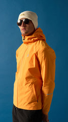 Person wearing an orange jacket, off-white OCL beanie with gold embroidery, and sunglasses against a blue background