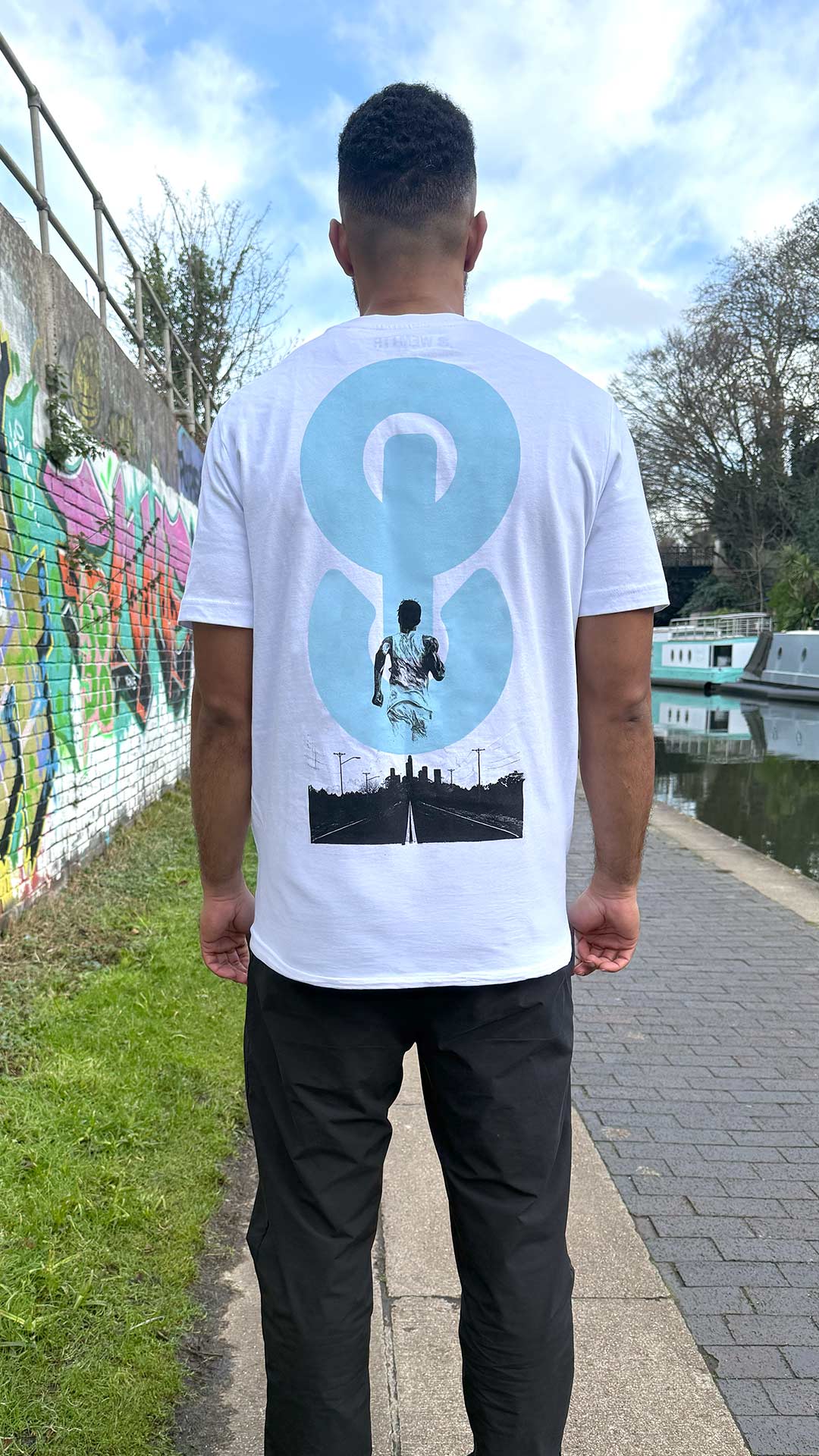 Person wearing a white t-shirt with a graphic design, standing on a path near a graffiti-covered wall.