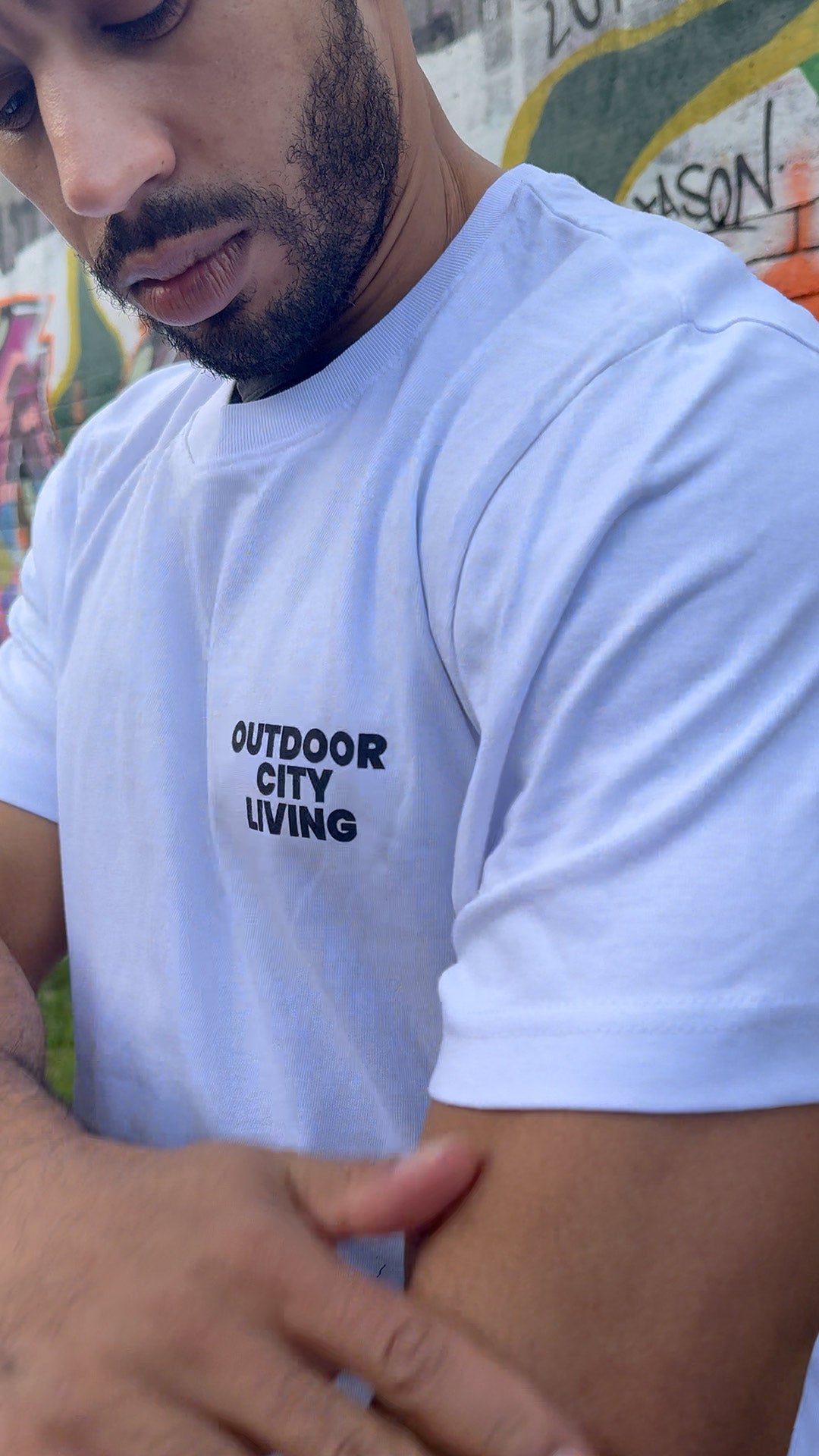 Person wearing a white t-shirt with 'Outdoor City Living' text, standing against a colorful mural.