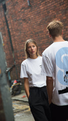 Two people wearing white t-shirts standing against a brick wall.